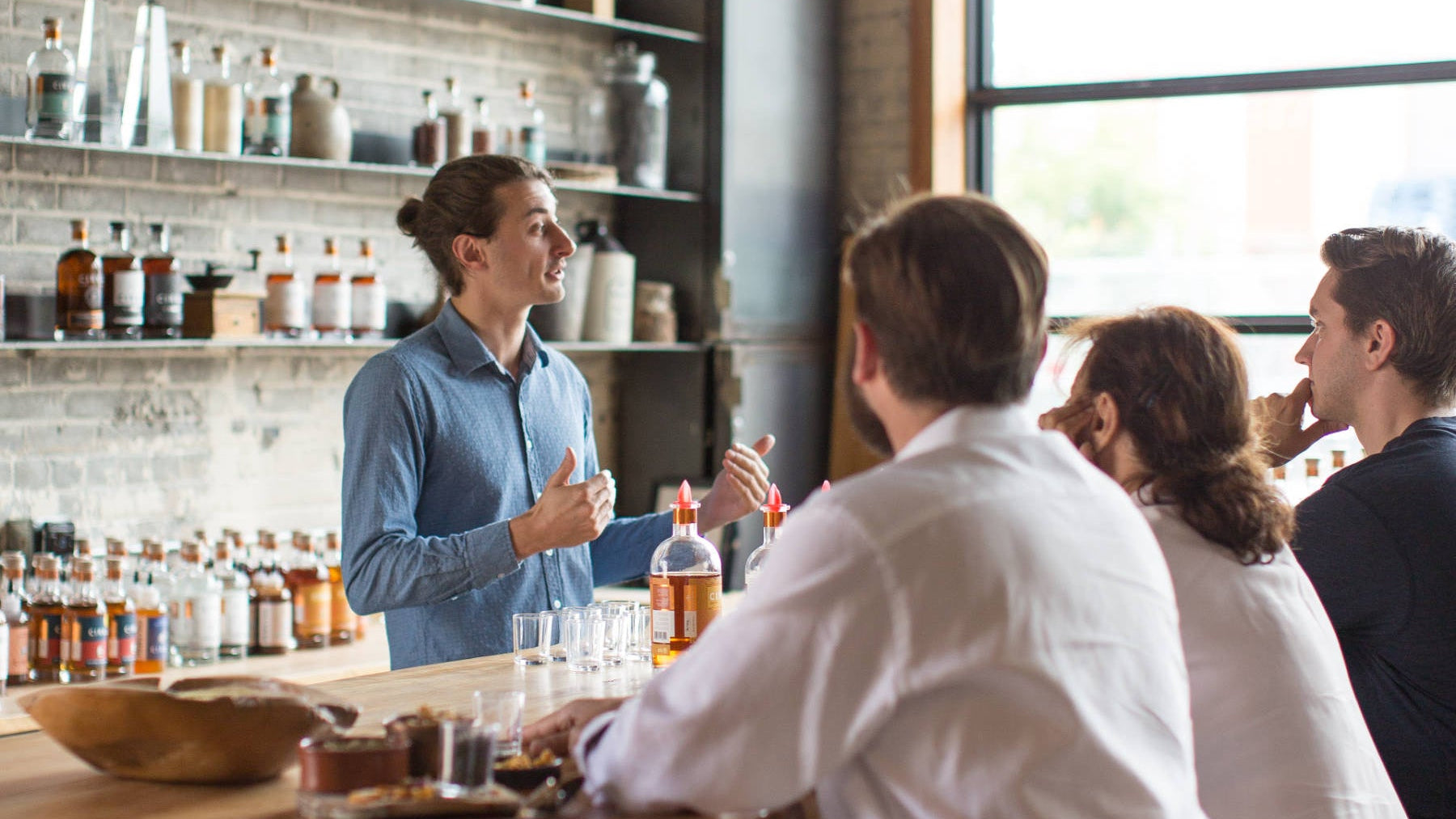 Des personnes assises au comptoir du bar de dégustation CIRKA en compagnie d'un barman, entourées d'étagères remplies de bouteilles et de grandes baies vitrées.